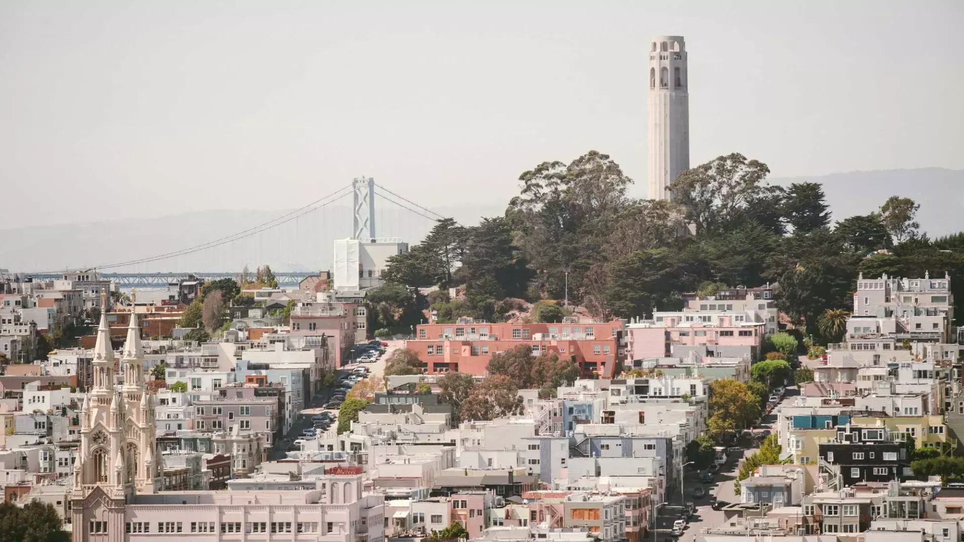 San Francisco's 屁股塔 is pictured with the Bay Bridge in the background 和 a hill covered in houses in the foreground.