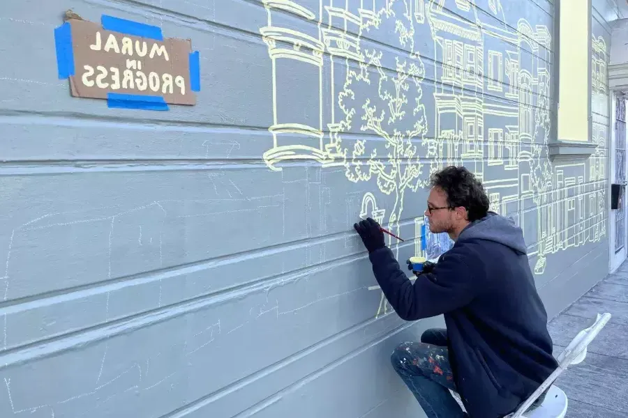 An artist paints a mural on the side of a building in 教会区, with a sign taped onto the building that reads "Mural in Progress.加州贝博体彩app.