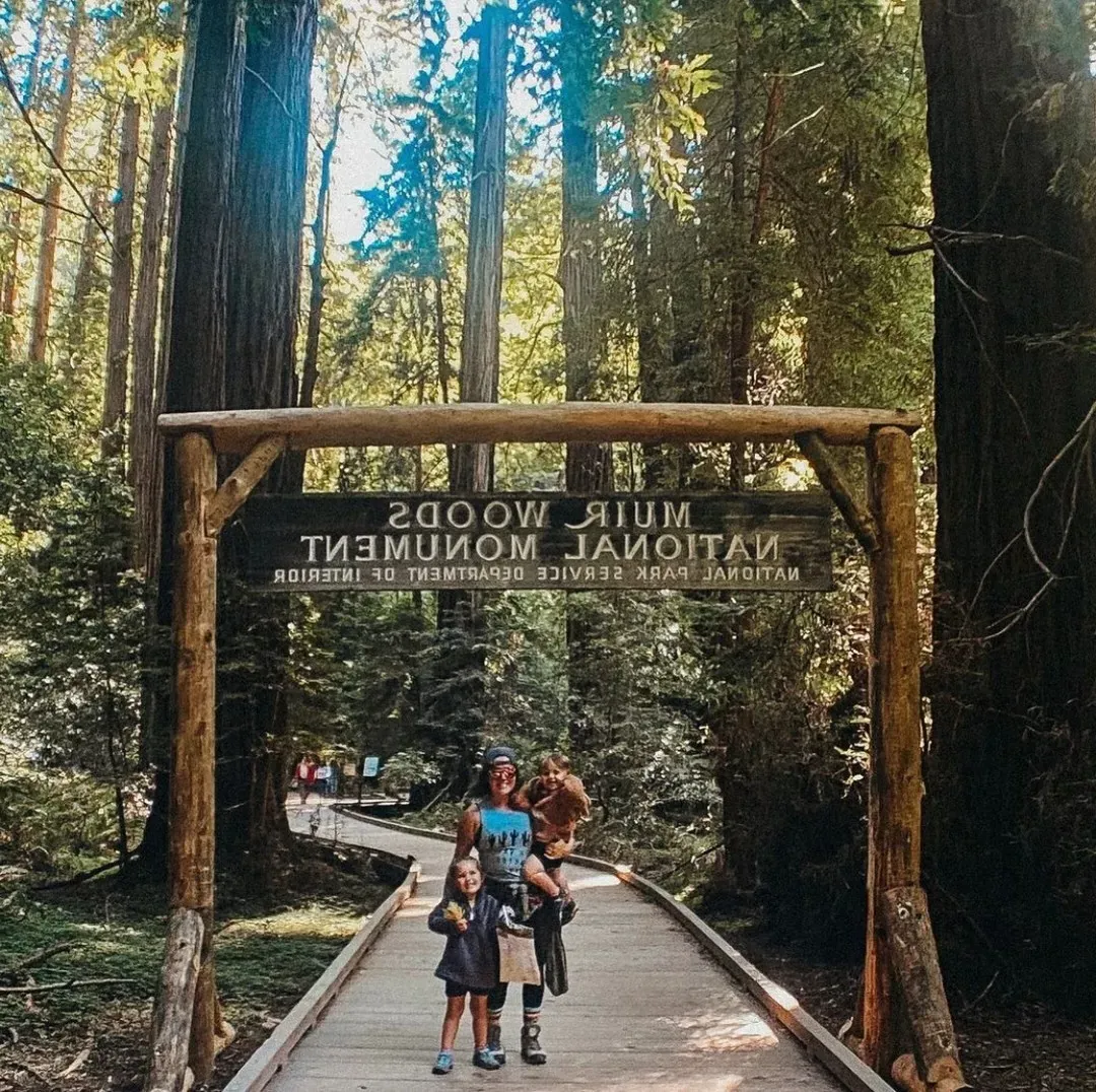 Woman and children stand in front of Muir Woods National Monument signage ona wooden trail in a forest